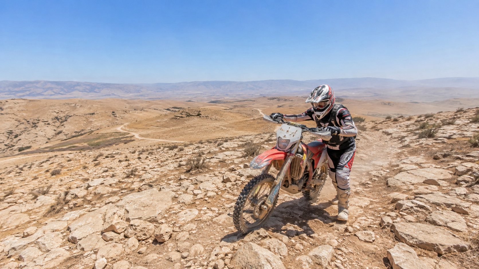 רוכב אנדורו דוחף אופנוע בעלייה סלעית תחת שמש צהריים, בלי צל בשום מקום